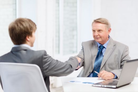 business, technology and office concept - older man and young man shaking hands in office