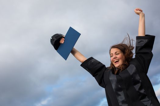 Pretty, young woman celebrating joyfully her graduation - spreading wide her arms, holding her diploma, savouring her success (color toned image; shallow DOF)