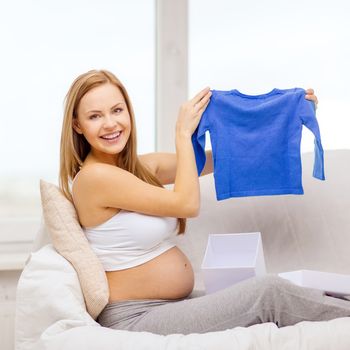 pregnancy, motherhood and celebration concept - smiling pregnant woman sitting on sofa and opening gift box with blue cardigan