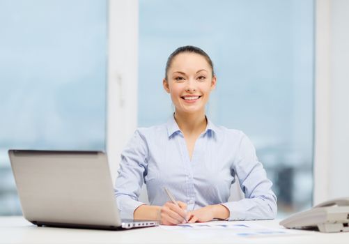 business, technology and communication concept - smiling businesswoman with phone, laptop and files in office