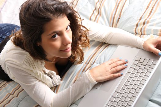 A young girl laying on the bed and surfing on the Internet with a Laptop.  