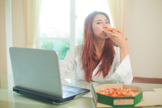 Happy asian young woman using laptop and eating pizza at home