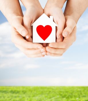 charity, real estate and family home concept - closeup picture of male and female hands holding white paper house with red heart