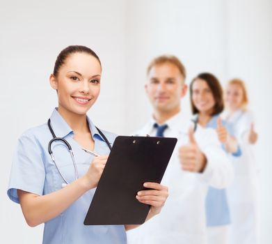 healthcare and medicine concept - smiling female doctor or nurse with stethoscope and clipboard with team on the back showing thumbs up