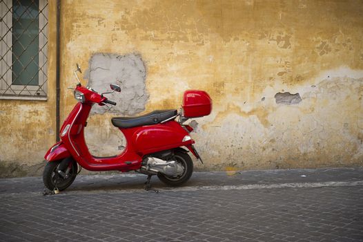 Bright shiny red Vespa scooter with a carrier parked on a sidewalk in front of a grungy dilapidated townhouse with peeling plaster on the wall and copyspace