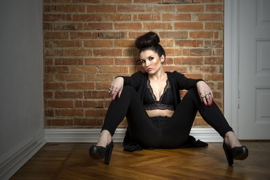 Moody beautiful elegant woman in a fashionable stylish black outfit sitting on a parquet floor with her legs spread looking at the camera with a sultry speculative expression