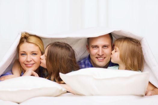 family, children and home concept - two little girls kissing their parents on cheek under blanket at home