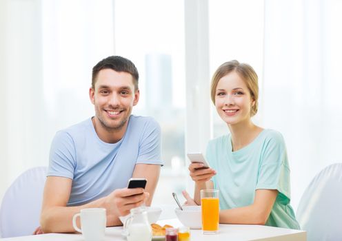 food, home, couple and technology concept - smiling couple with smartphones reading news and having breakfast at home