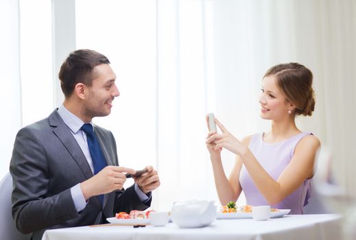 restaurant, couple, technology and holiday concept - smiling woman taking picture of husband or boyfriend while picturing sushi