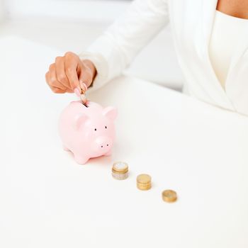 business and money saving concept - close up of female hand putting euro coins into piggy bank in office