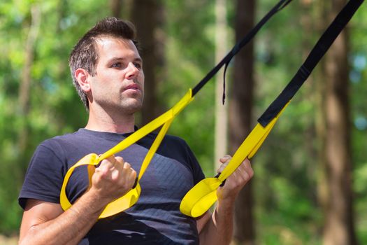Young attractiveman does suspension training with fitness straps outdoors in the nature.