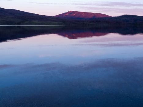 Reflection of distant sunset boreal forest taiga mountain mirrored on calm water surface of Little Salmon Lake, Yukon Territory, Canada