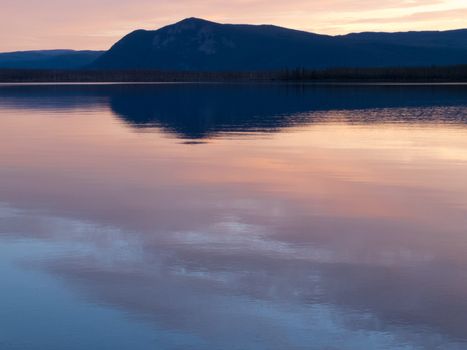 Reflection of distant sunset boreal forest taiga mountain mirrored on calm water surface of Little Salmon Lake, Yukon Territory, Canada