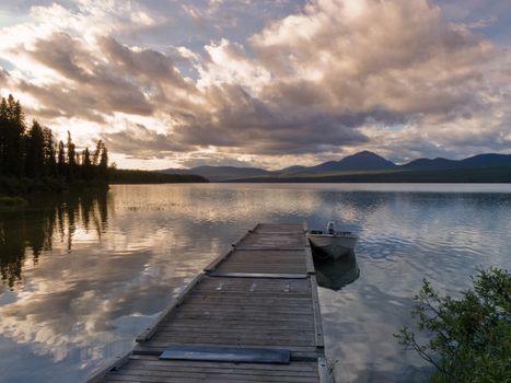 Rustic wooden floating dock or jetty on a tranquil lake, small john boat moored and dramatic clouds sunset reflected on still water