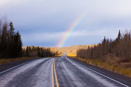 Wet asphalt of North Klondike Highway road leading to colourful rainbow over late autumn fall forested landscape after rain in Yukon Territory, Canada