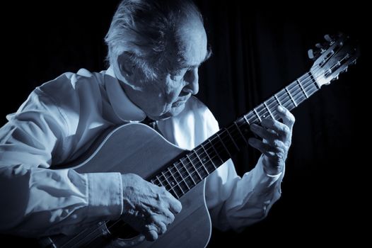 An elderly man in white shirt playing an acoustic guitar. Dark background. Monochrome.