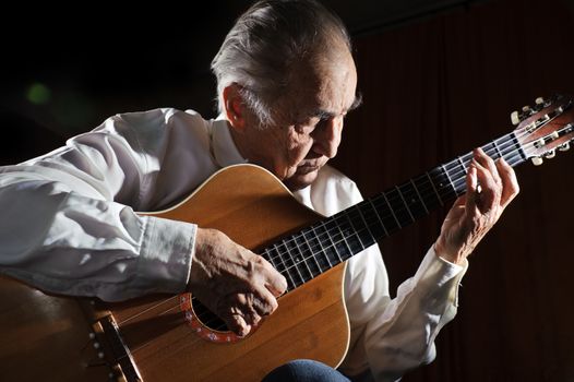 An elderly man in white shirt playing an acoustic guitar. Dark background. 