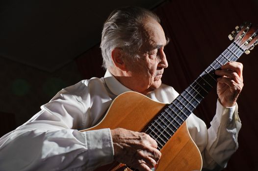 An elderly man in white shirt playing an acoustic guitar. Dark background. 