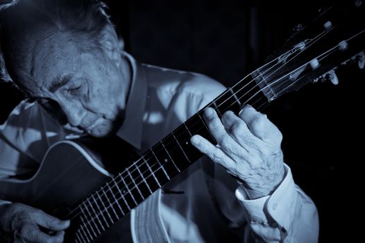 An elderly man in white shirt playing an acoustic guitar. Dark background. Monochrome.  Focus on the hand.