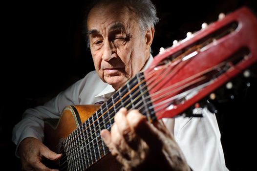 An elderly man in white shirt playing an acoustic guitar. Dark background. 