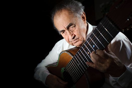 An elderly man in white shirt playing an acoustic guitar. Dark background. 