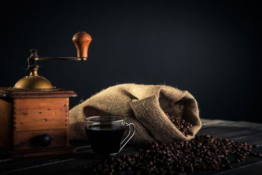 tools for preparation of coffee. an old grinder a sack made by jute full of coffee beans and cup of black coffee on a wooden table.