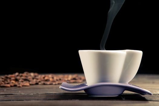 close up of a steaming cup of coffee with coffee beans scattered behind on a wooden table and a black background
