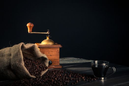 tools for preparation of coffee. an old grinder a sack made by jute full of coffee beans and cup of black coffee on a wooden table.