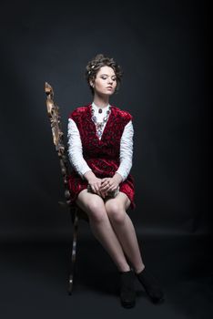 Portrait of beautiful Caucasian model in red Victorian-style velvet dress, seated on wooden chair