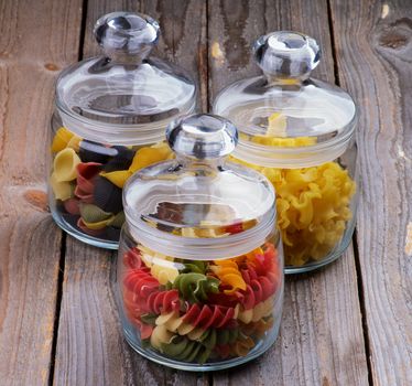 Three Glass Jars with Colorful Conchiglie Pasta, Ballerine Pasta and Rotini Pasta isolated on Wooden background