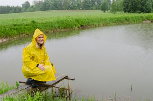 girl sits on the foot bridge to the village pond on rainy day with yellow rain coat