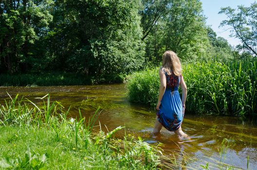 blond girl with long blue mottled summer dress wade through fast flowing river in summer day