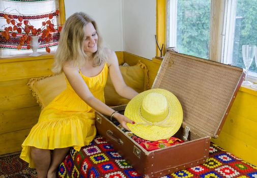 smiling woman put nice summer yellow hat in old vintage suitcase in village room