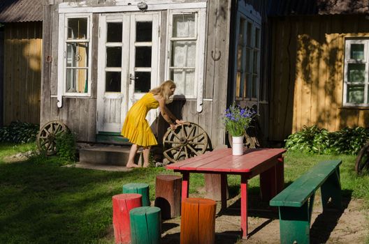 Rural wooden house yard and woman girl in yellow dress rolls old carriage wheel. Beautiful retro decoration. Colorful chairs and table and cornflowers bunch in vase.