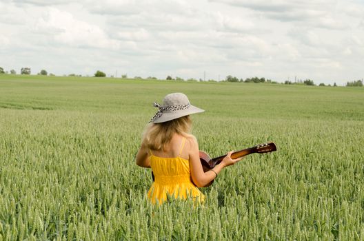 Country girl in yellow dress and hat on head play with guitar at wheat field.