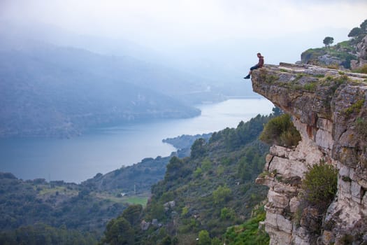 Young man sitting on edge of cliff and looking at river below