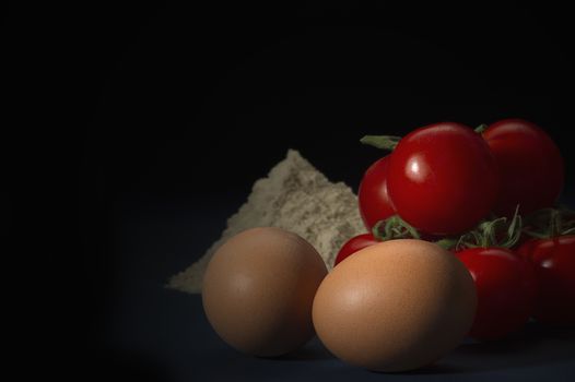 Fresh brown eggs, cherry tomatoes and flour standing ready on a kitchen counter to be cooked or used as ingredients in baking, dark background with copyspace