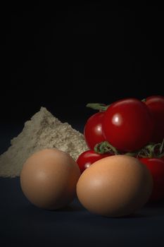 Healthy fresh ingredients including brown free range eggs, flour and ripe cherry tomatoes in a country kitchen ready to be used in cooking, vertical format on a dark background with copyspace