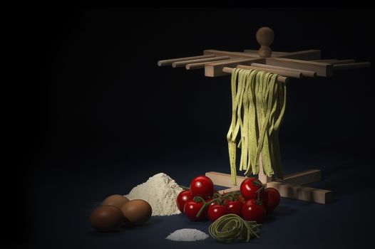 Ingredients for preparing a traditional Italian pasta with taglitelli hanging on a wooden frame, cherry tomatoes, flour, eggs and parmesan cheese on a kitchen counter, dark background with copyspace