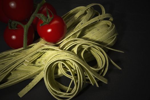 Coiled dried tagliatelle or fettuccine Italian pasta with a bunch of fresh ripe red cherry tomatoes ready to be used in preparing Italian cuisine on a dark background
