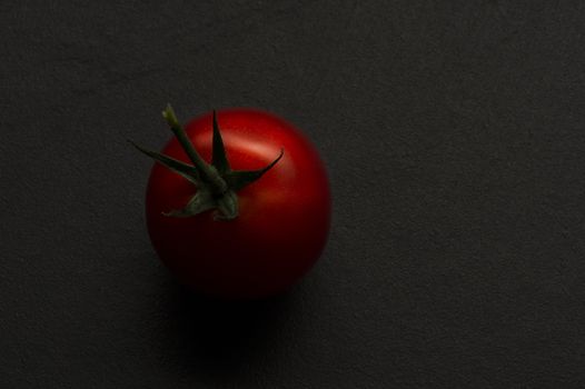 Single fresh whole organic ripe red hothouse tomato with a green stalk on a dark background with copyspace