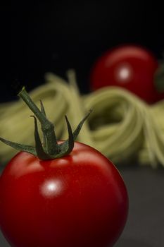 Luscious ripe red fresh tomato with an attached green stalk with Italian pasta ready to cook a traditional Mediterranean meal, vertical format on a dark background with copyspace