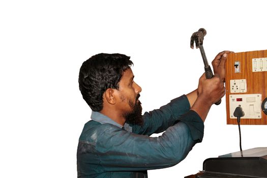 Profile waist level view of a casually dressed , bearded Indian handy man in dusty green shirt , seen working  on a switchboard with a raised hammer in the right hand appears to be in an office setting and the whole scene is isolated on white background