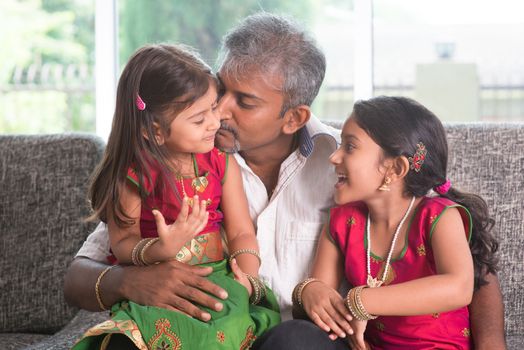 Happy Indian family at home. Asian father kissing her daughter, sitting on sofa. Parent and children indoor lifestyle.