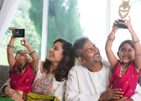 Happy Asian Indian family at home. Kid raised their trophy high up, education achievement concept. Parents and children indoor lifestyle.
