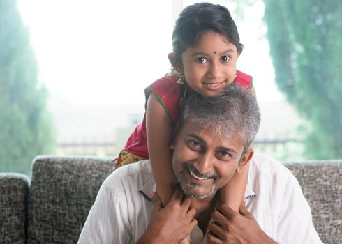 Happy Indian family at home. Asian father piggyback her daughter, sitting on sofa. Parent and child indoor lifestyle.