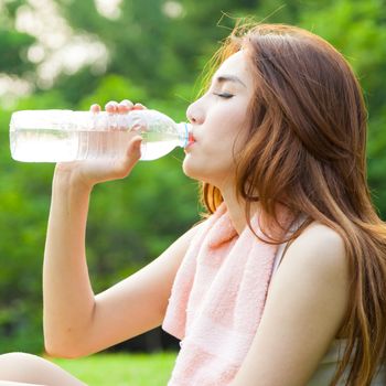 Woman sitting tired and drinking water after exercise. Within the lawn of park.