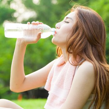 Woman sitting tired and drinking water after exercise. Within the lawn of park.