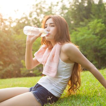 Woman sitting tired and drinking water after exercise. Within the lawn of park.