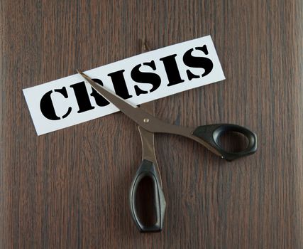 Scissors cutting the word "Crisis" written on a paper strip, over wooden background